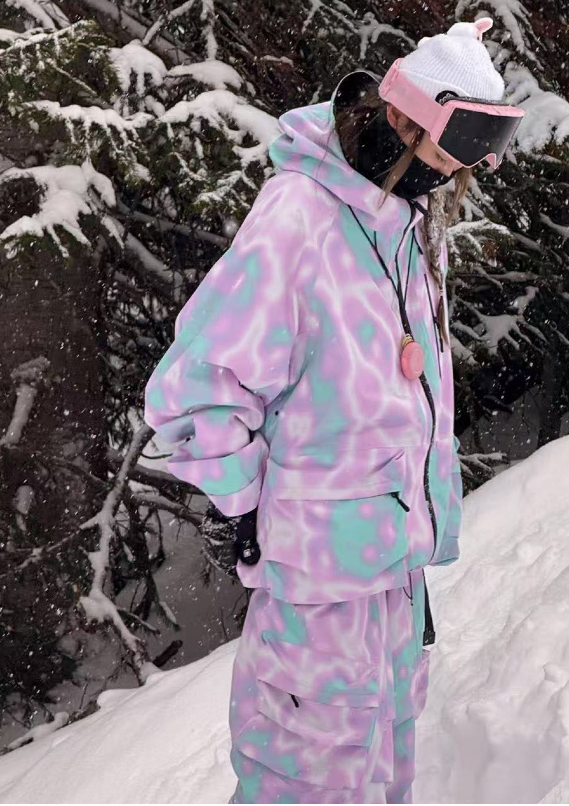 Person wearing a colorful snowsuit with a pink helmet and goggles in a snowy forest.